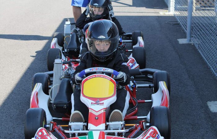Deux enfants portant des casques sont assis dans des karts sur la piste pour enfants de Go-kart Mont-Tremblant, alignés sur une piste asphaltée et se préparant à faire la course, avec une personne se tenant à proximité.