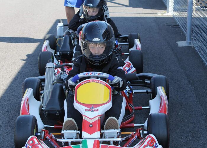 Two children wearing helmets and racing suits sit in go-karts on a paved track, preparing to drive.