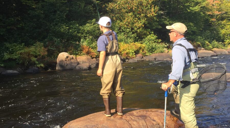 Two people in waders stand on a rock by a river, fishing with rods and nets, surrounded by trees and greenery. Two people in waders stand on a rock by a river, fishing with rods and nets, surrounded by trees and greenery.