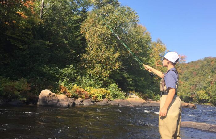 Person wearing waders and a cap is fishing with a rod in a river surrounded by rocks and trees on a sunny day.