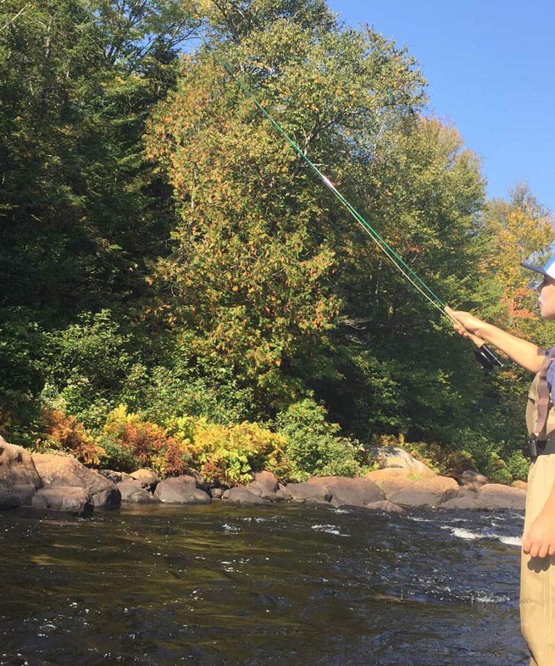 Une personne portant des cuissardes et une casquette se tient dans une rivière, profitant d'une découverte en famille pendant la Pêche à la mouche, entourée d'arbres et de rochers par une journée ensoleillée.