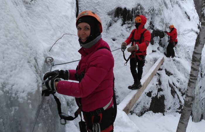 Three people in winter gear are climbing a snowy, icy mountainside using safety ropes and harnesses.