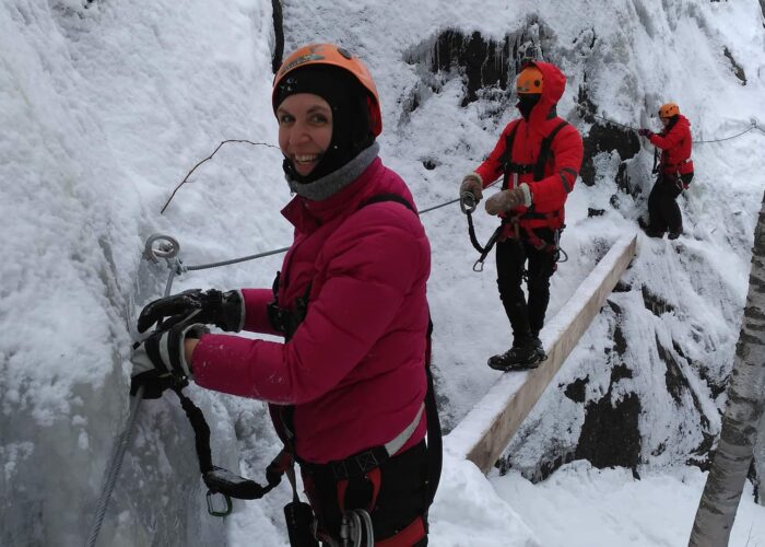 Three people in winter gear are climbing a snowy, icy mountainside using safety ropes and harnesses.