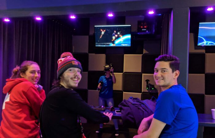 Three people sit at a counter watching someone wearing a VR headset play a space-themed game, with monitors displaying the gameplay above.
