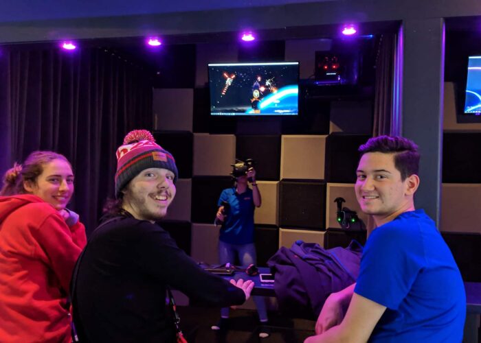 Three people sit at a counter watching someone wearing a VR headset play a space-themed game, with monitors displaying the gameplay above.