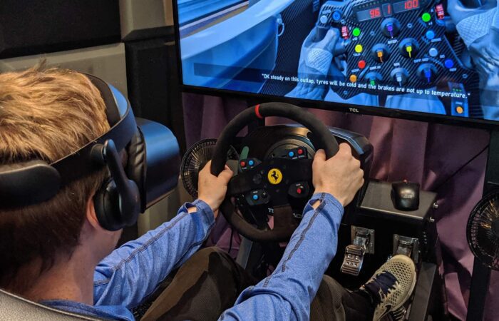 A person wearing a VR headset uses a racing simulator with a steering wheel and pedals, looking at a large screen displaying a racing game.
