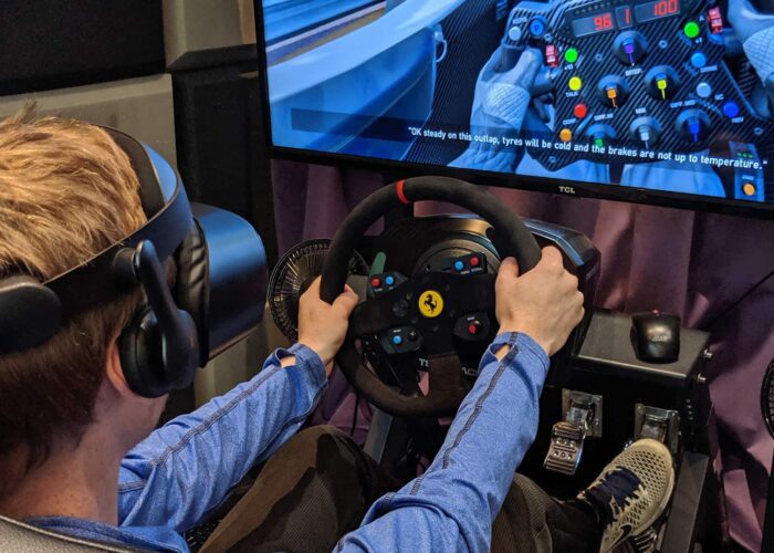 A person wearing a VR headset uses a racing simulator with a steering wheel and pedals, looking at a large screen displaying a racing game.