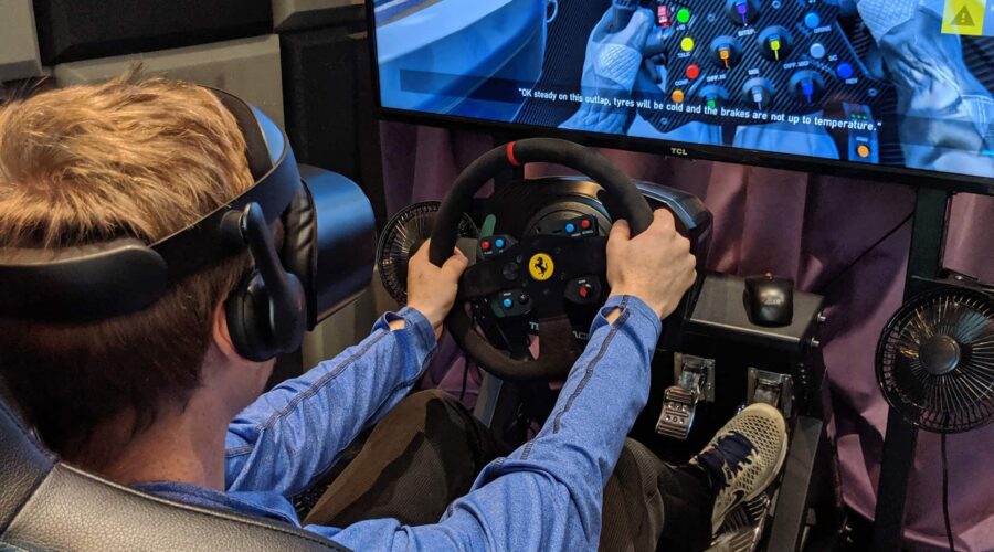 Person wearing a VR headset uses a racing wheel controller while playing a car racing simulation game on a large monitor.