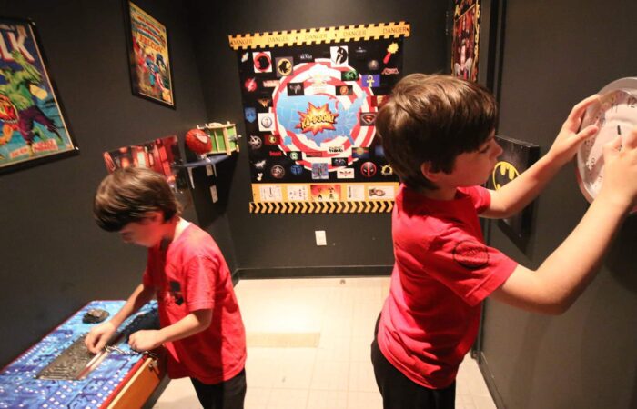 Two boys in red shirts solve puzzles in a superhero-themed escape room decorated with posters and props related to comic book characters. Two boys in red shirts solve puzzles in a superhero-themed escape room decorated with posters and props related to comic book characters.