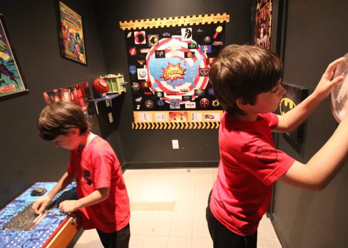 Two boys in red shirts solve puzzles in a superhero-themed escape room decorated with posters and props related to comic book characters.