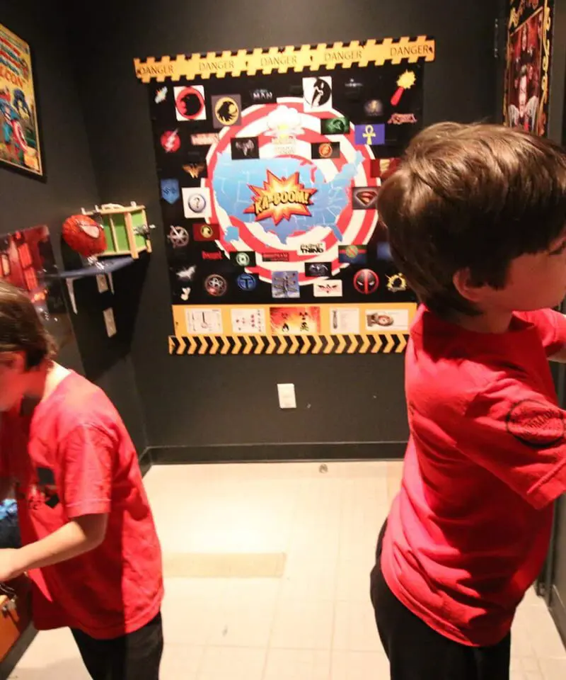 Two boys in red shirts solve puzzles in a superhero-themed escape room decorated with posters and props related to comic book characters.