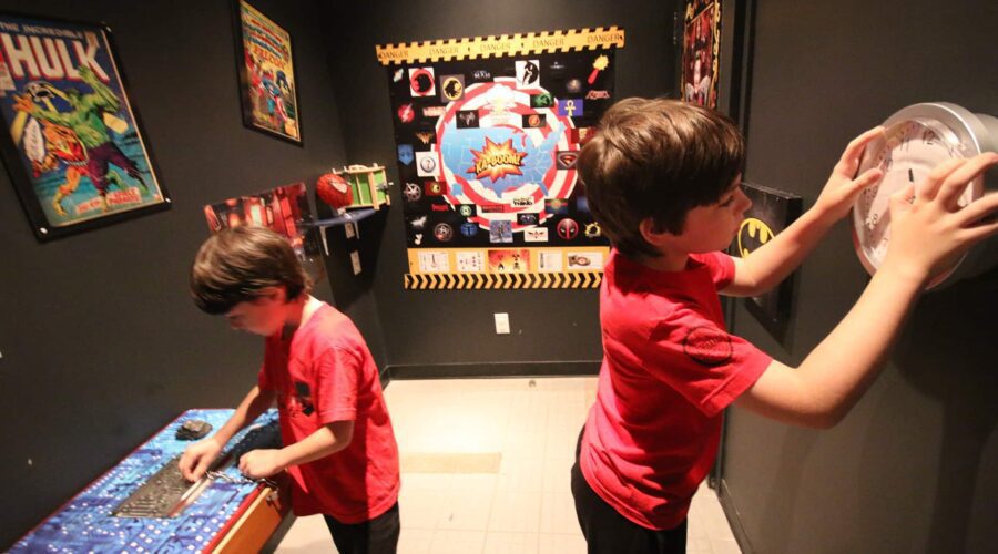 Two boys in red shirts solve puzzles in a superhero-themed escape room decorated with posters and props related to comic book characters.
