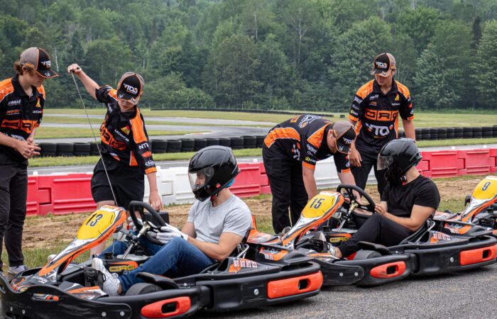 Plusieurs personnes en uniforme de course préparent des karts pour trois pilotes portant des casques, assis dans des karts numérotés sur une piste régulière à Mont-Tremblant, avec des arbres bordant la piste extérieure en arrière-plan.