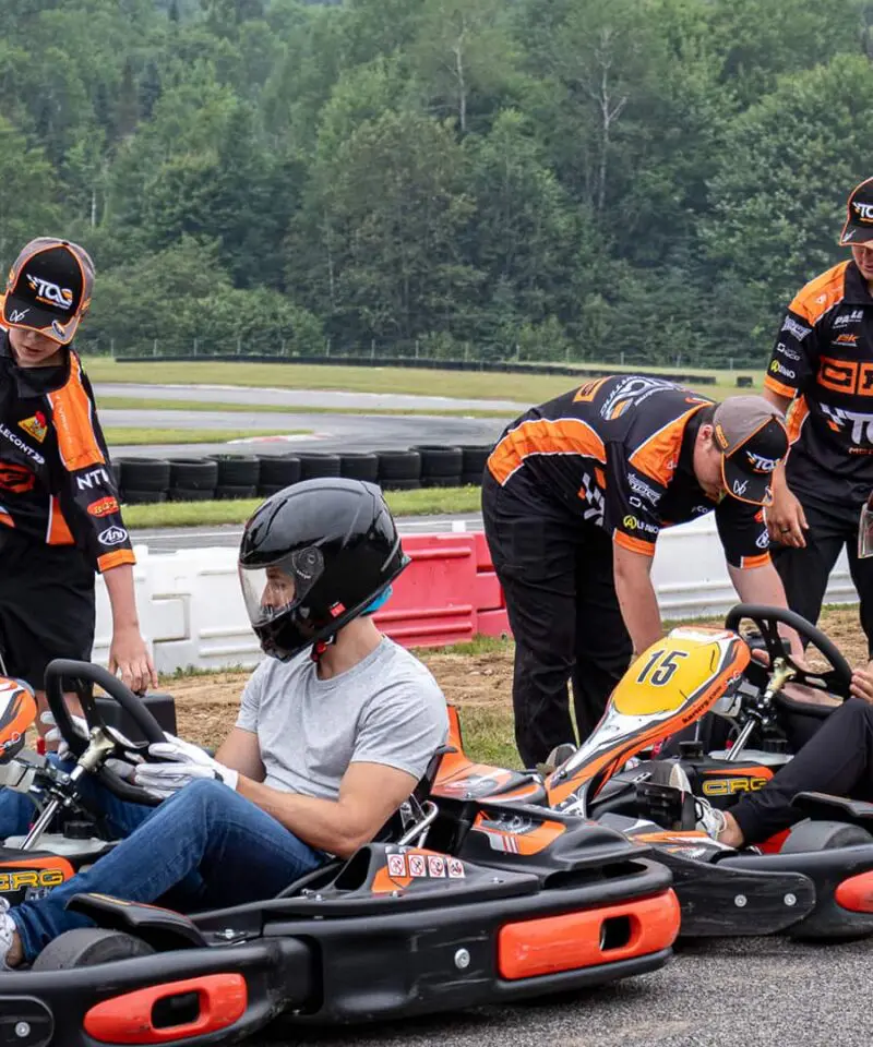 Three people sit in go-karts wearing helmets while three team members in matching uniforms stand nearby, preparing the karts on an outdoor track. Trees and barriers are visible in the background.