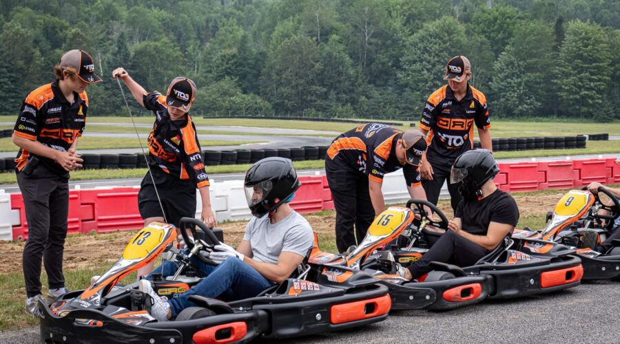 Three people sit in go-karts wearing helmets while three team members in matching uniforms stand nearby, preparing the karts on an outdoor track. Trees and barriers are visible in the background. Three people sit in go-karts wearing helmets while three team members in matching uniforms stand nearby, preparing the karts on an outdoor track. Trees and barriers are visible in the background.