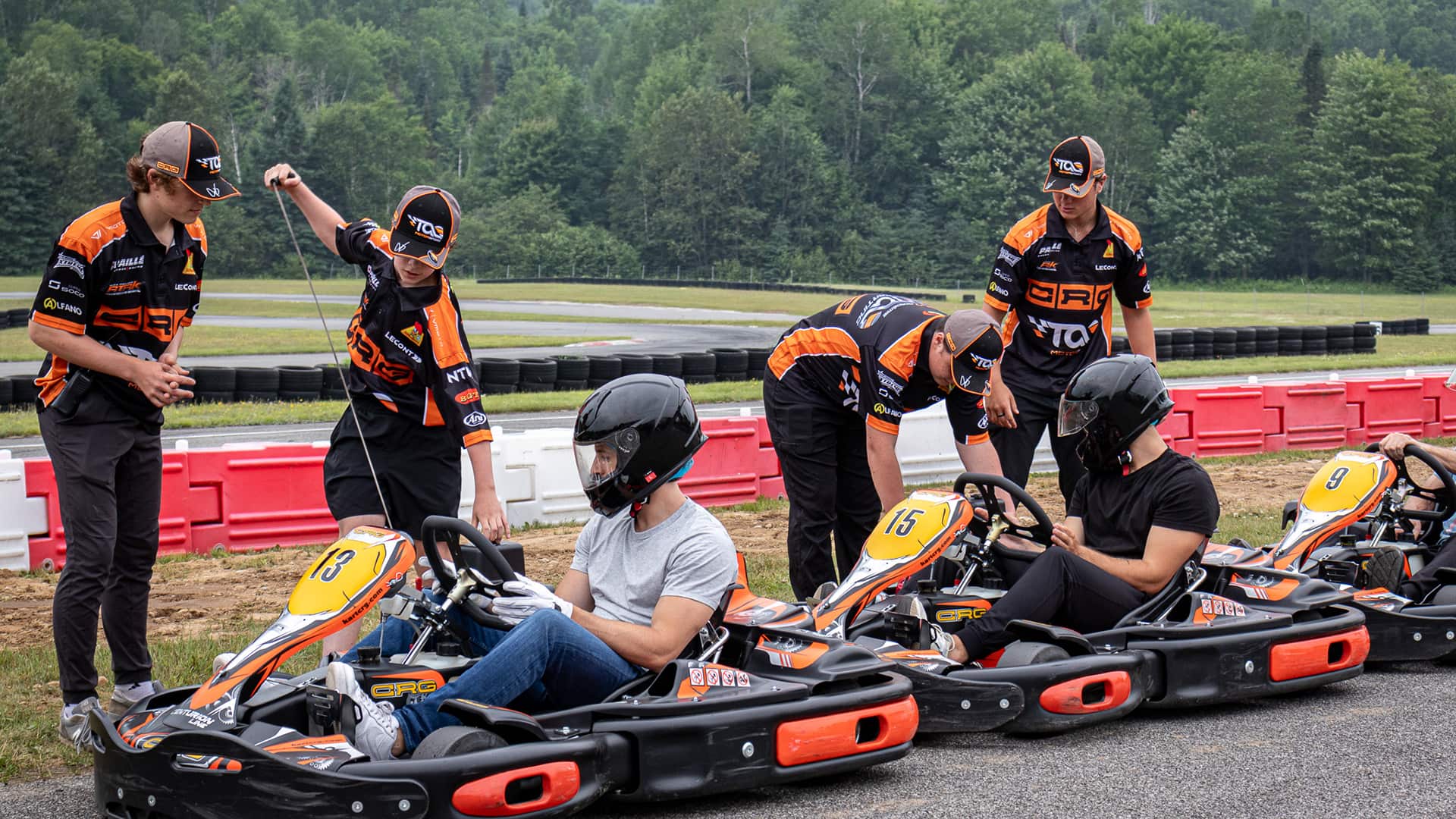 Three people sit in go-karts wearing helmets while three team members in matching uniforms stand nearby, preparing the karts on an outdoor track. Trees and barriers are visible in the background.