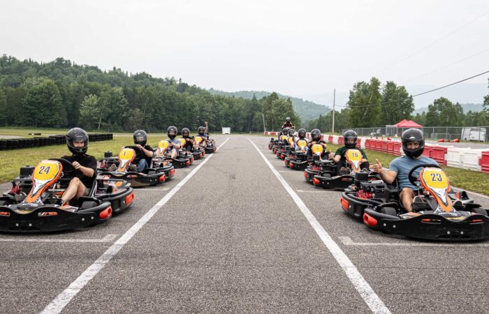 Un groupe de personnes portant des casques est assis dans des karts alignés sur une piste extérieure à Mont-Tremblant, prêts à faire la course. Des arbres et des collines sont visibles à l'arrière-plan, plantant un décor pittoresque pour ces courses privées palpitantes.