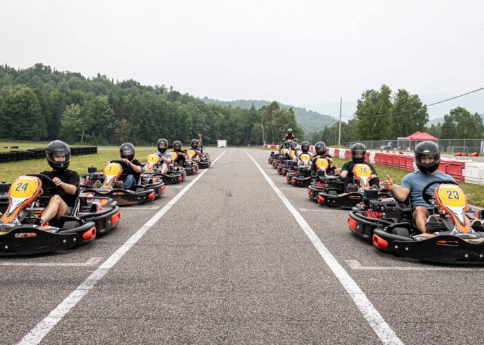 A group of people wearing helmets sit in go-karts lined up on an outdoor racetrack, ready to start. Trees and hills are visible in the background.