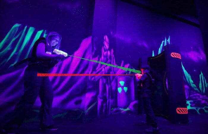 Two people play laser tag in a dark, neon-lit arena with glowing vests and laser beams.