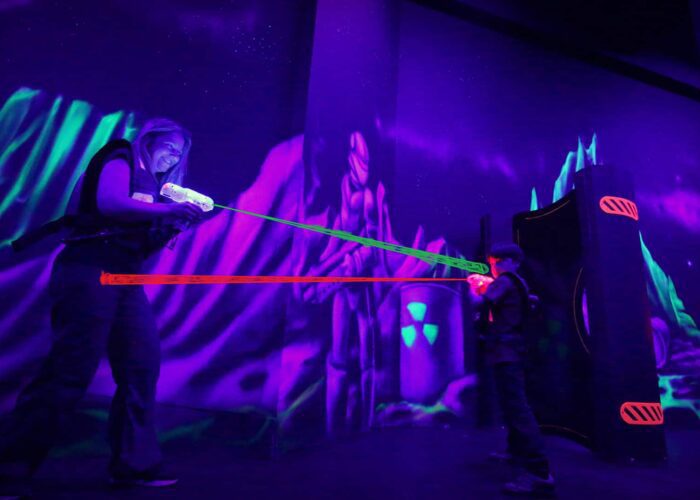 Two people play laser tag in a dark, neon-lit arena with glowing vests and laser beams.