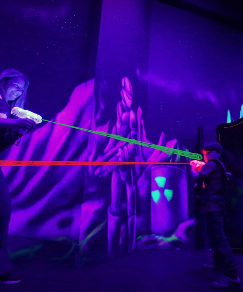 Two people play laser tag in a dark, neon-lit arena with glowing vests and laser beams.