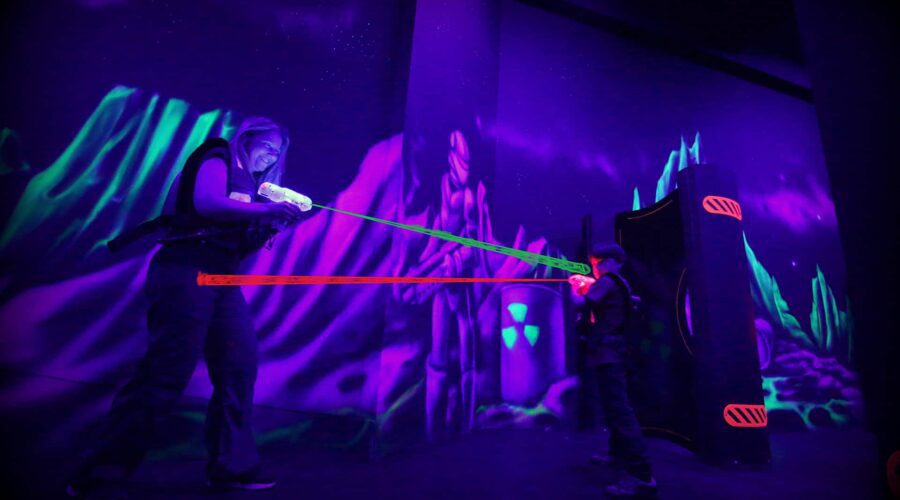 Two people play laser tag in a dark, neon-lit arena with glowing vests and laser beams.