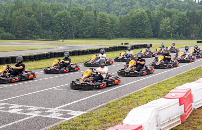 A group of people wearing helmets sit in go-karts lined up at the starting line of an outdoor race track surrounded by greenery. A group of people wearing helmets sit in go-karts lined up at the starting line of an outdoor race track surrounded by greenery.