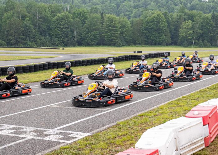 A group of people wearing helmets sit in go-karts lined up at the starting line of an outdoor race track surrounded by greenery.