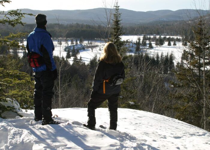 Two people in winter clothing stand on a snowy hill overlooking a snow-covered landscape with trees and distant hills.