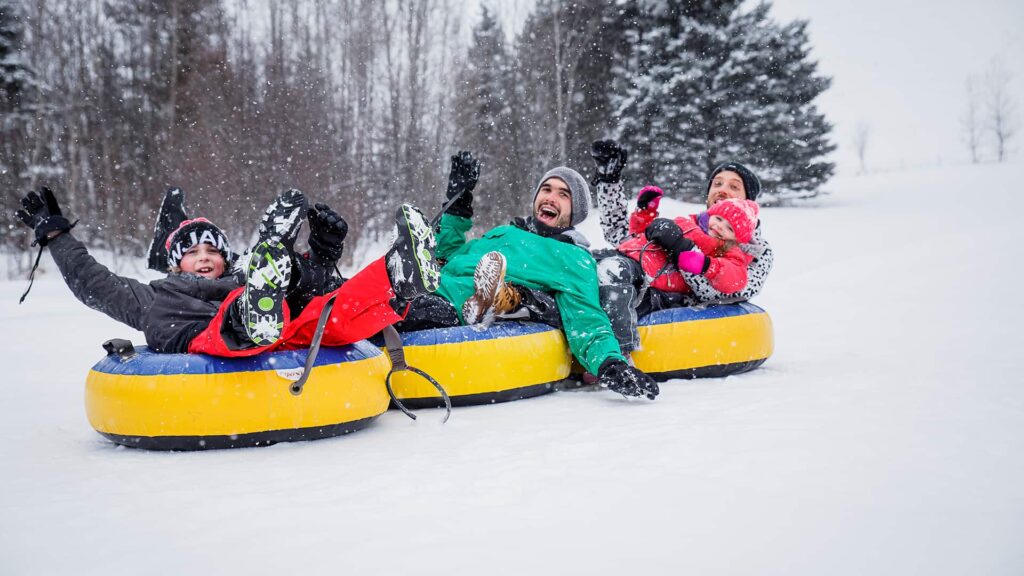 Three people and a child ride two yellow snow tubes down a snowy hill, surrounded by trees, on a winter day.