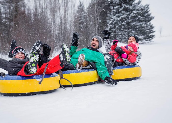 Three people and a child ride two yellow snow tubes down a snowy hill, surrounded by trees, on a winter day. Three people and a child ride two yellow snow tubes down a snowy hill, surrounded by trees, on a winter day.