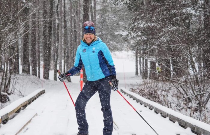 Person cross-country skiing on a snow-covered path through a forest, wearing a blue jacket, winter hat, gloves, and sunglasses, with snow falling heavily.