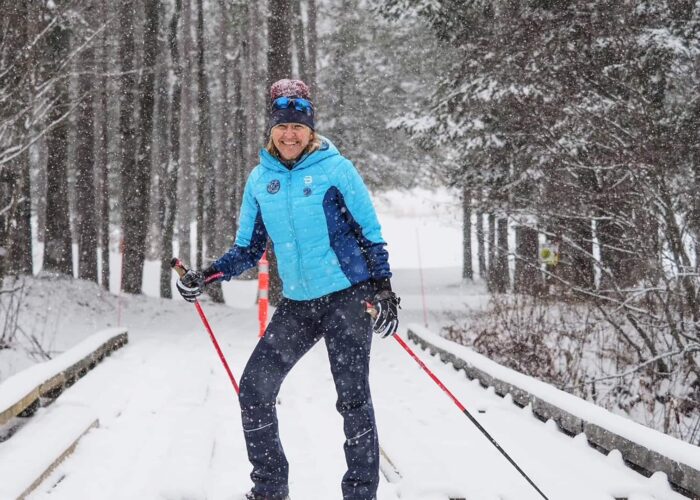 Person cross-country skiing on a snow-covered path through a forest, wearing a blue jacket, winter hat, gloves, and sunglasses, with snow falling heavily.