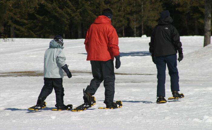 Three people wearing winter jackets and snowshoes walk across a snowy landscape with trees in the background.