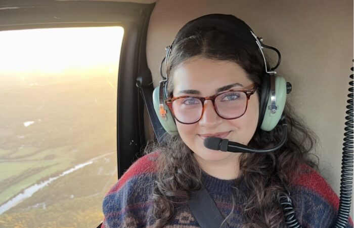 A woman wearing glasses and a headset sits inside a helicopter, smiling at the camera with a landscape visible through the window.