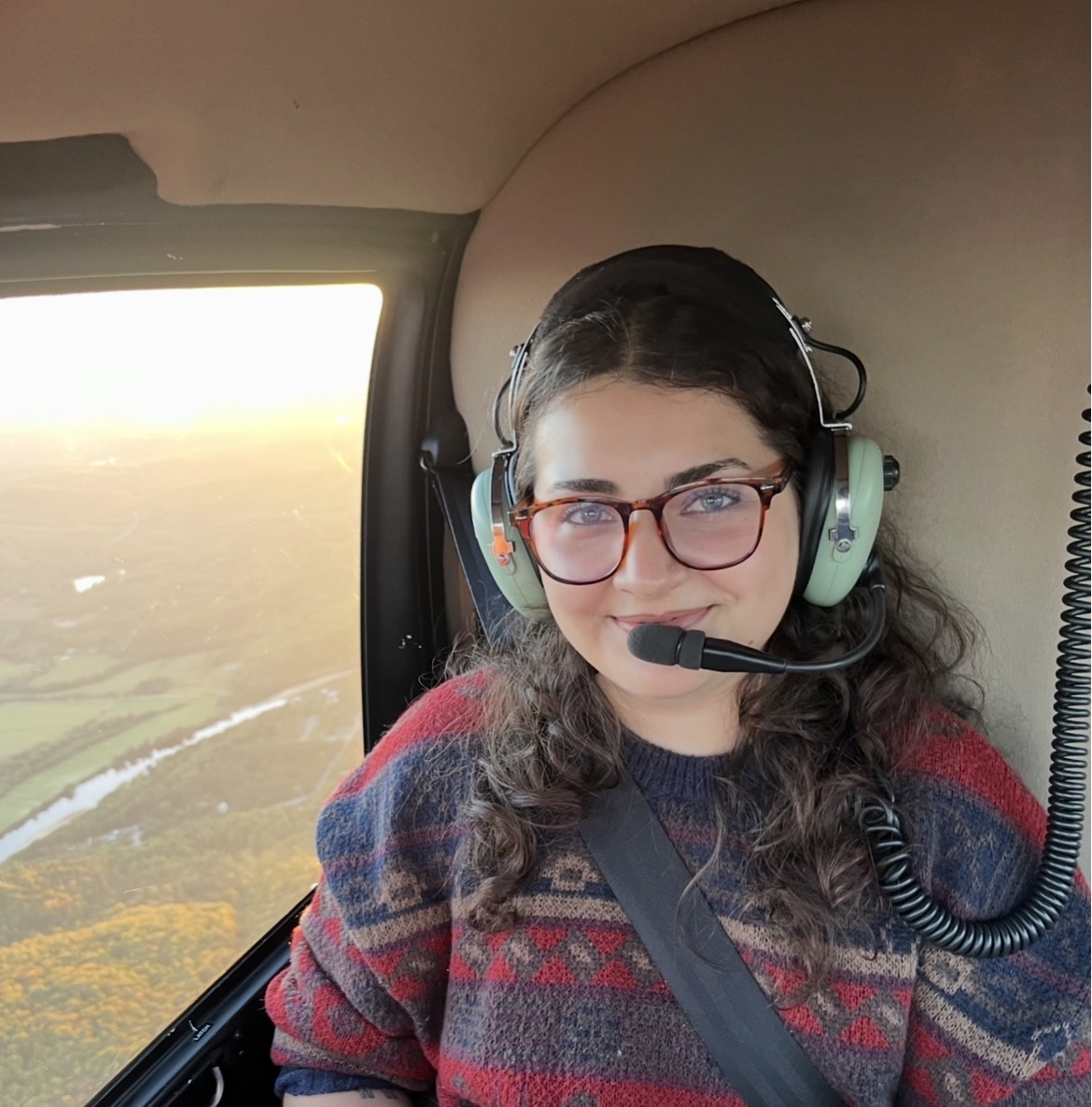 A woman wearing glasses and a headset sits inside a helicopter, smiling at the camera with a landscape visible through the window. A woman wearing glasses and a headset sits inside a helicopter, smiling at the camera with a landscape visible through the window.
