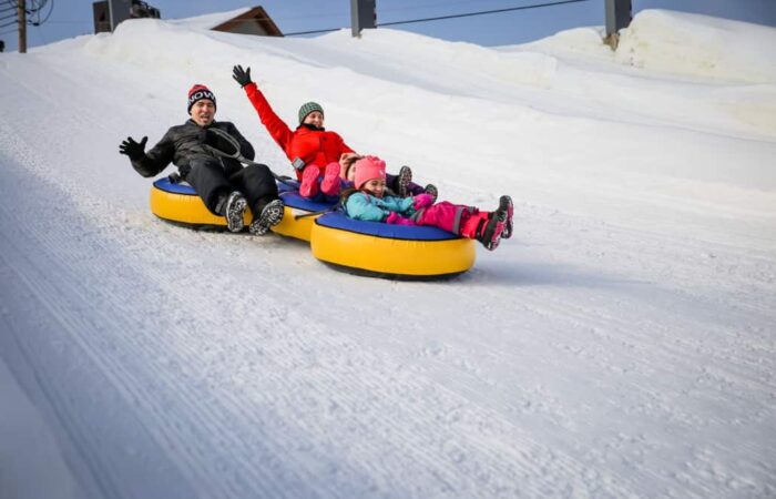 Four people, two adults and two children, ride yellow snow tubes down a snowy hill, wearing winter clothing and appearing excited.
