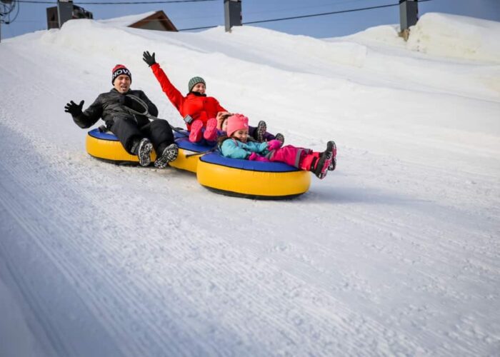Four people, two adults and two children, ride yellow snow tubes down a snowy hill, wearing winter clothing and appearing excited.