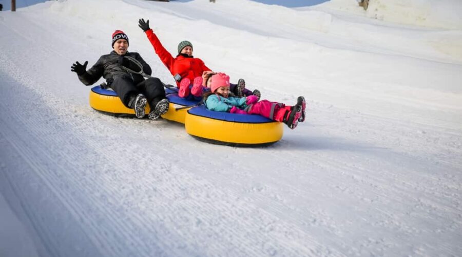Four people, two adults and two children, ride yellow snow tubes down a snowy hill, wearing winter clothing and appearing excited.