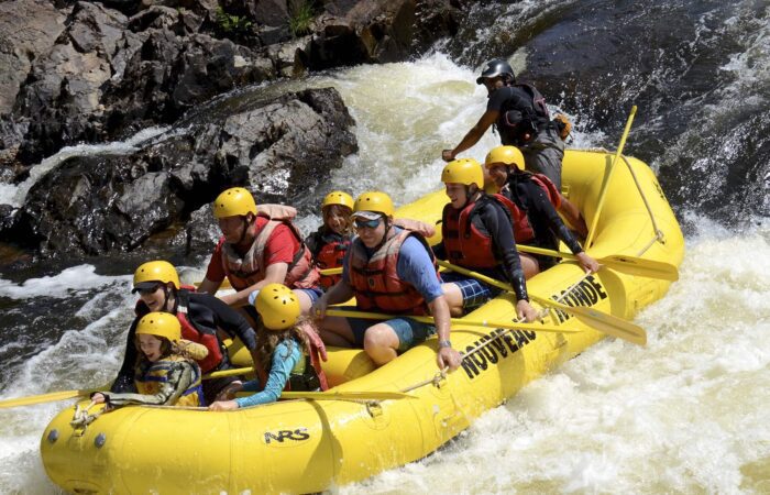 A group of people wearing helmets and life jackets paddle a yellow inflatable raft down a rocky, whitewater river.