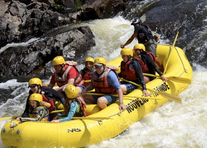 A group of people wearing helmets and life jackets paddle a yellow inflatable raft down a rocky, whitewater river.