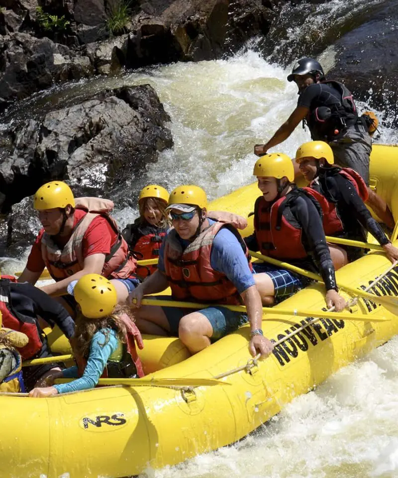 A group of people wearing helmets and life jackets paddle a yellow inflatable raft down a rocky, whitewater river.