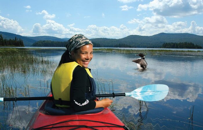 A person in a red kayak with a blue paddle and yellow life jacket smiles on a calm lake, with mountains and a bird on the water in the background.