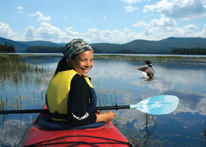 A person in a red kayak with a blue paddle and yellow life jacket smiles on a calm lake, with mountains and a bird on the water in the background.
