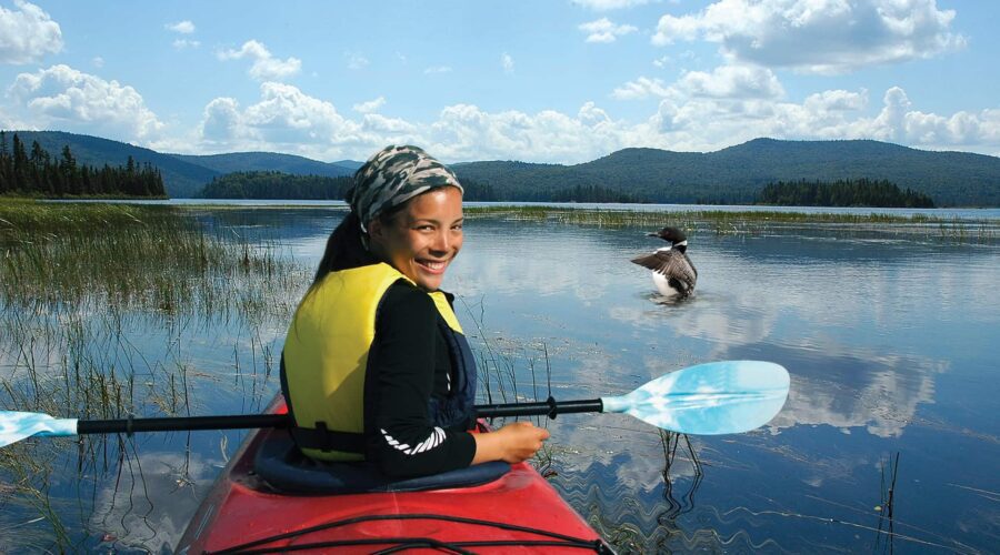 A person in a red kayak with a blue paddle and yellow life jacket smiles on a calm lake, with mountains and a bird on the water in the background.