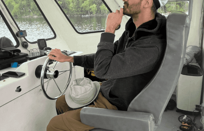 A man wearing glasses and a white captain's hat sits at the helm of a boat, holding the wheel with one hand and resting his chin on his other hand, looking forward.