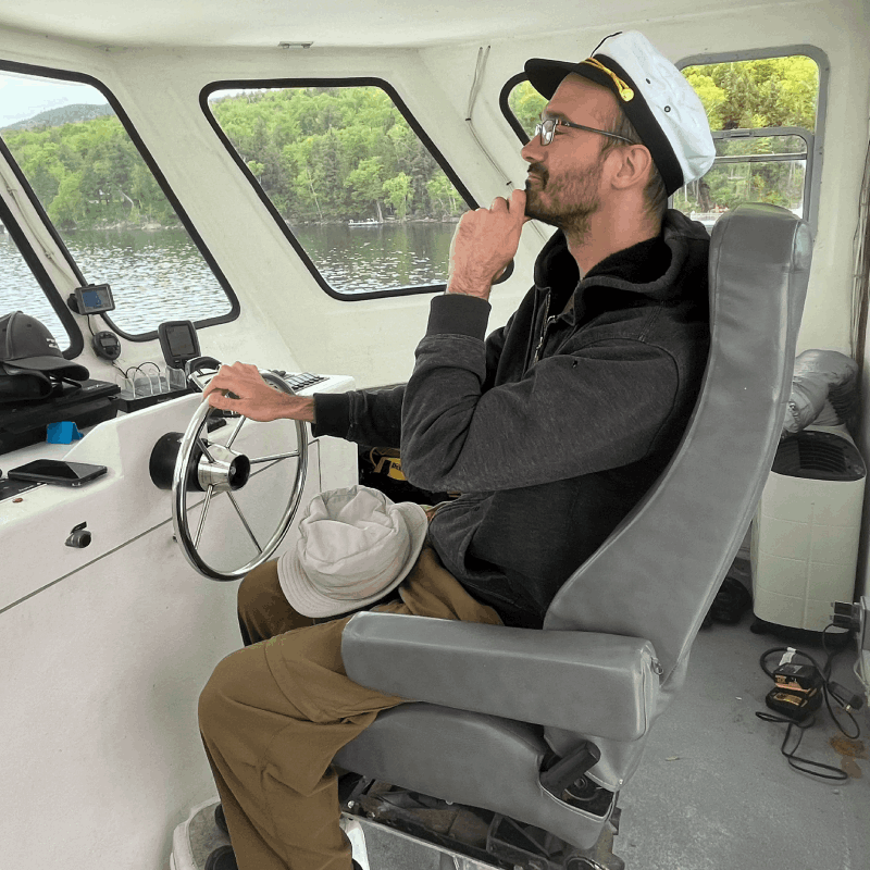 A man wearing glasses and a white captain’s hat sits at the helm of a boat, holding the wheel with one hand and resting his chin on his other hand, looking forward. A man wearing glasses and a white captain's hat sits at the helm of a boat, holding the wheel with one hand and resting his chin on his other hand, looking forward.