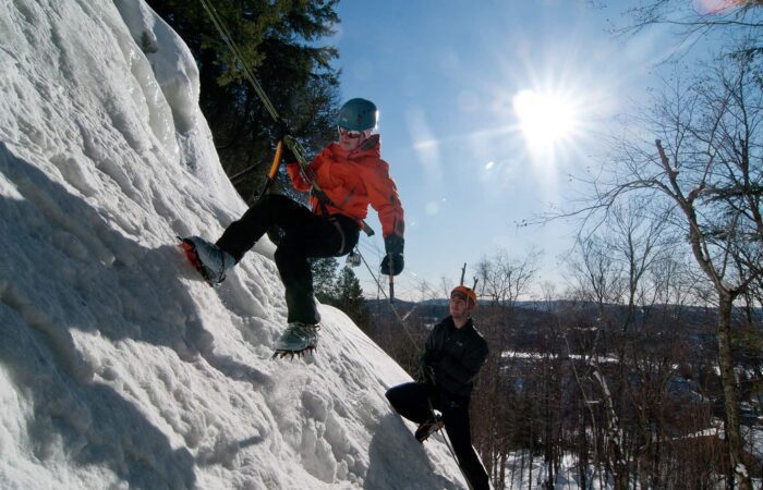 Two people ice climbing on a snowy slope; one climbs with gear while the other stands below holding a safety rope. The sun is shining in a clear sky.