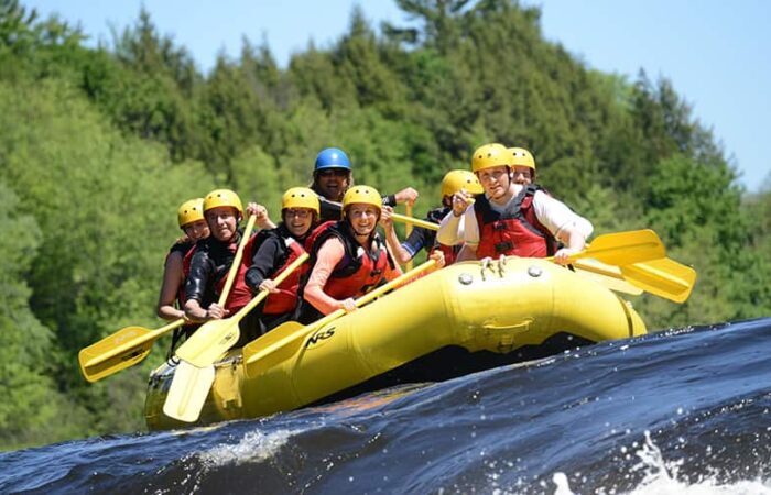 A group of people wearing helmets and life jackets paddle a yellow inflatable raft on a river surrounded by trees. A group of people wearing helmets and life jackets paddle a yellow inflatable raft on a river surrounded by trees.