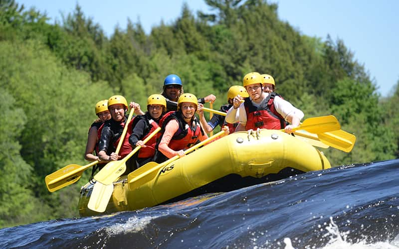 A group of people wearing helmets and life jackets paddle a yellow inflatable raft on a river surrounded by trees. A group of people wearing helmets and life jackets paddle a yellow inflatable raft on a river surrounded by trees.